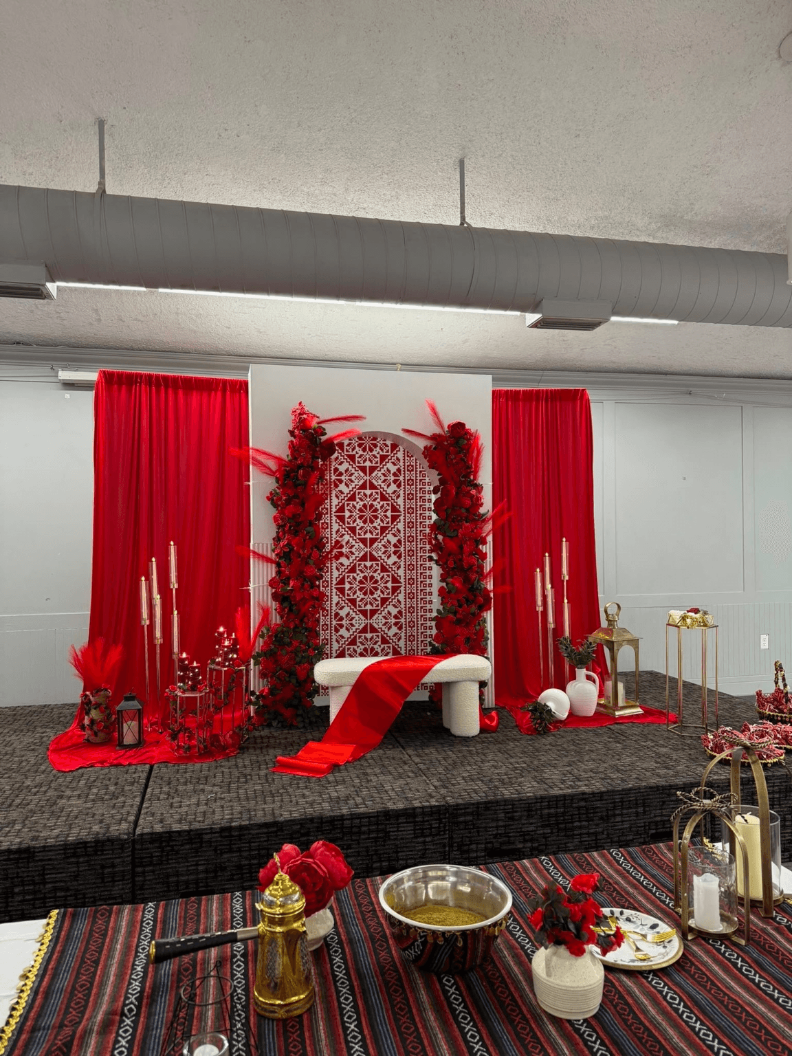 Floral stage with red drapery, red roses, a patterned arch, a white bench, and gold candleholders styled by Star Events